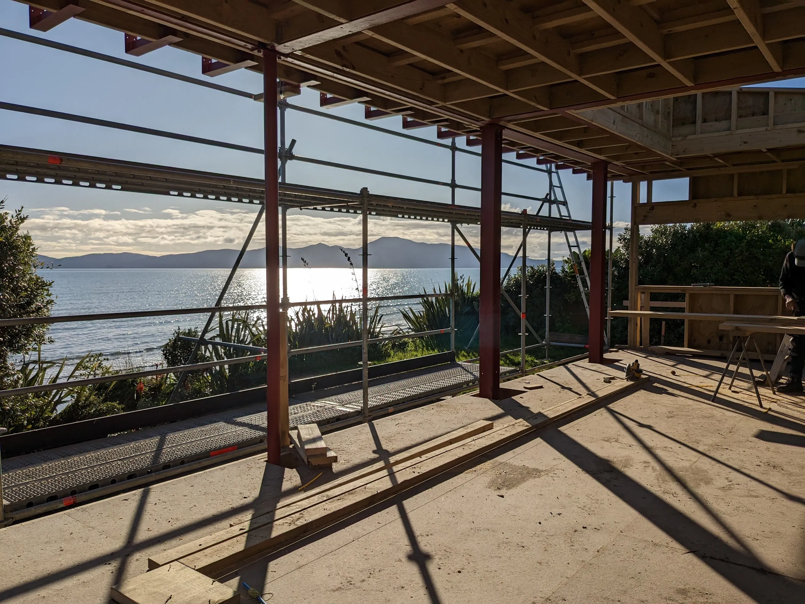Construction photograph from inside the house, looking past red primed steel columns and scaffolding to the sea and a large island beyond.