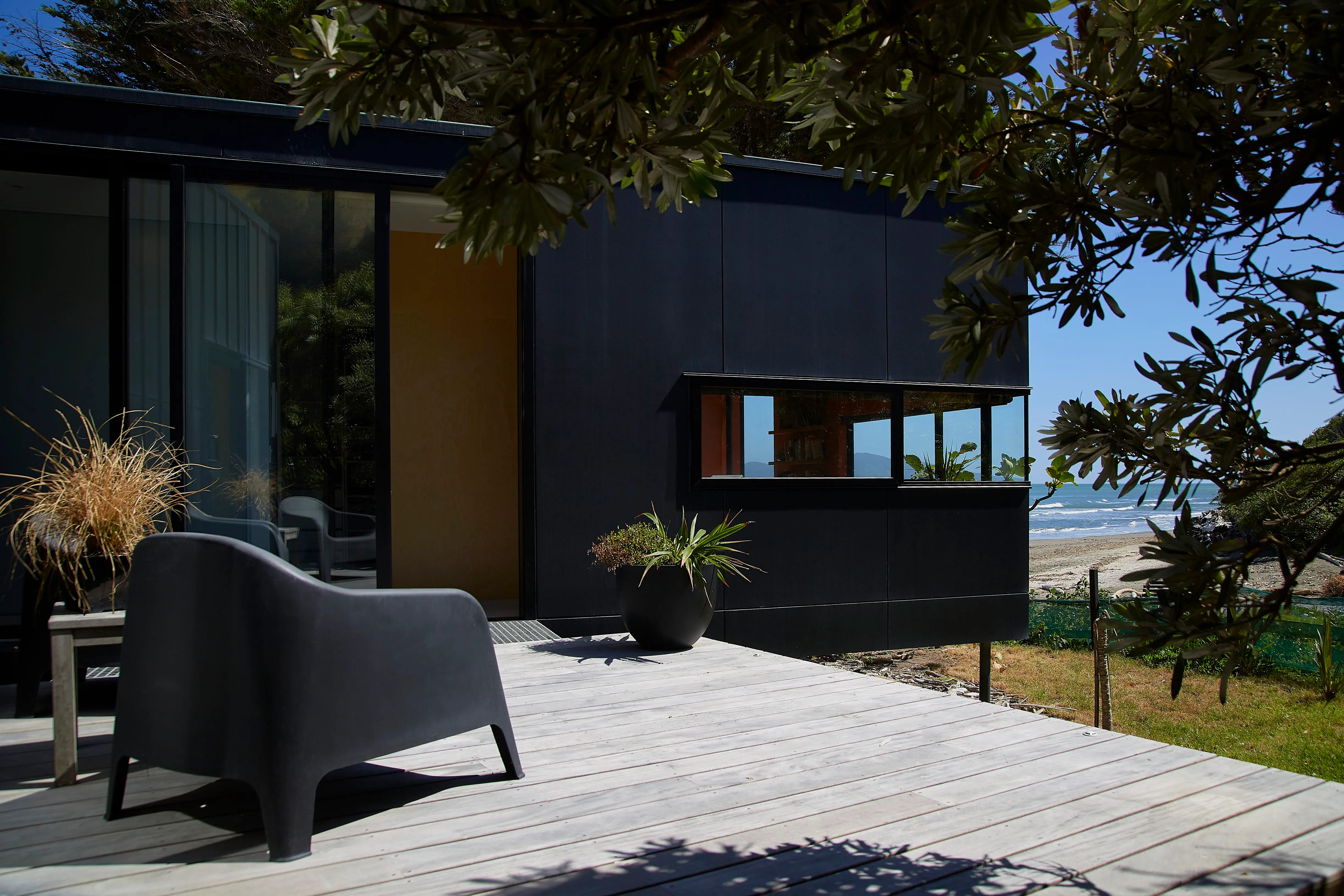 Timber deck beside the black-clad exterior of the house, with a long horizontal window and the beach and sea beyond.