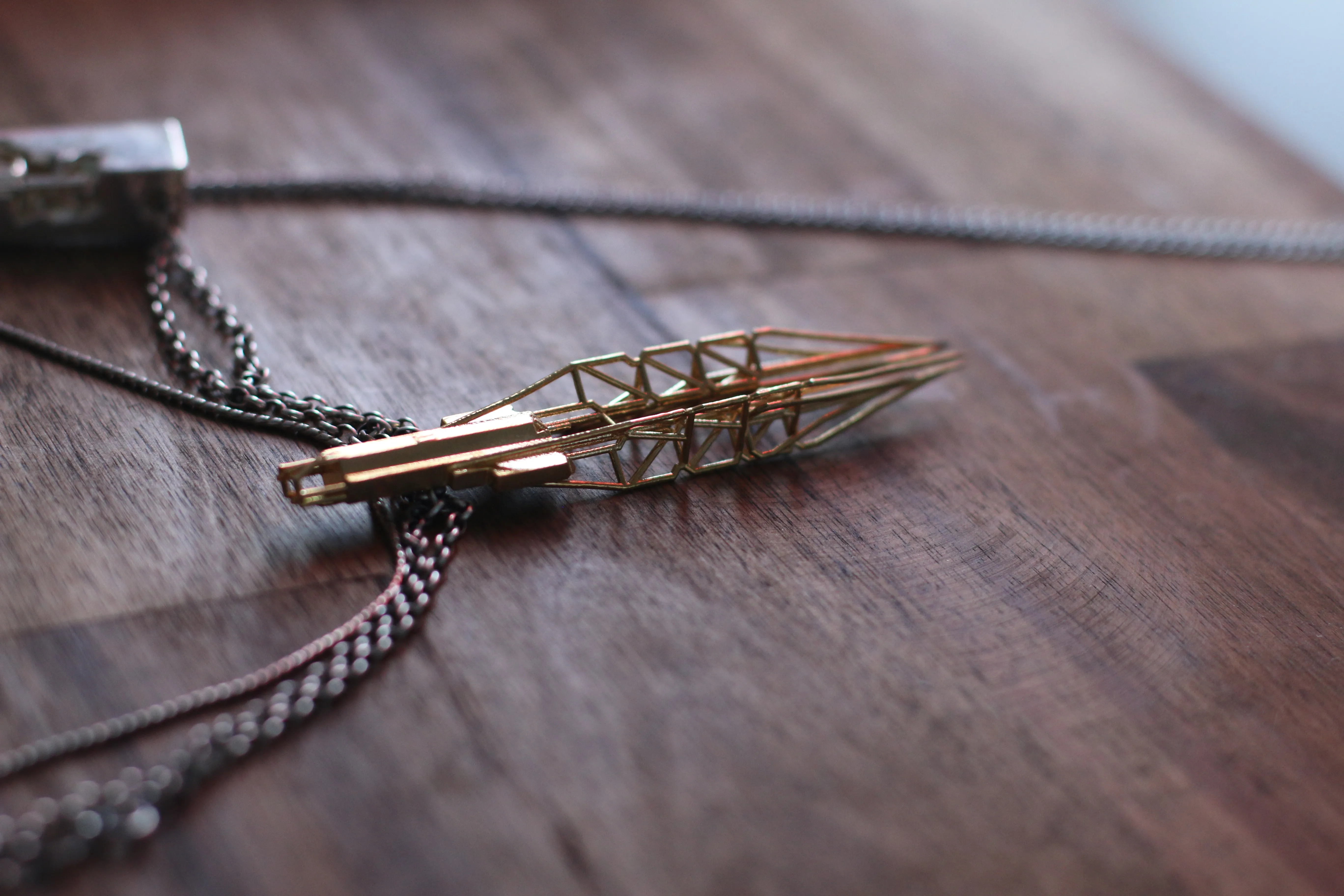 A gold-toned pendant on a dark chain, formed as a slender tapering spire of open trusswork rising from a solid squared base, resting on a dark timber table.