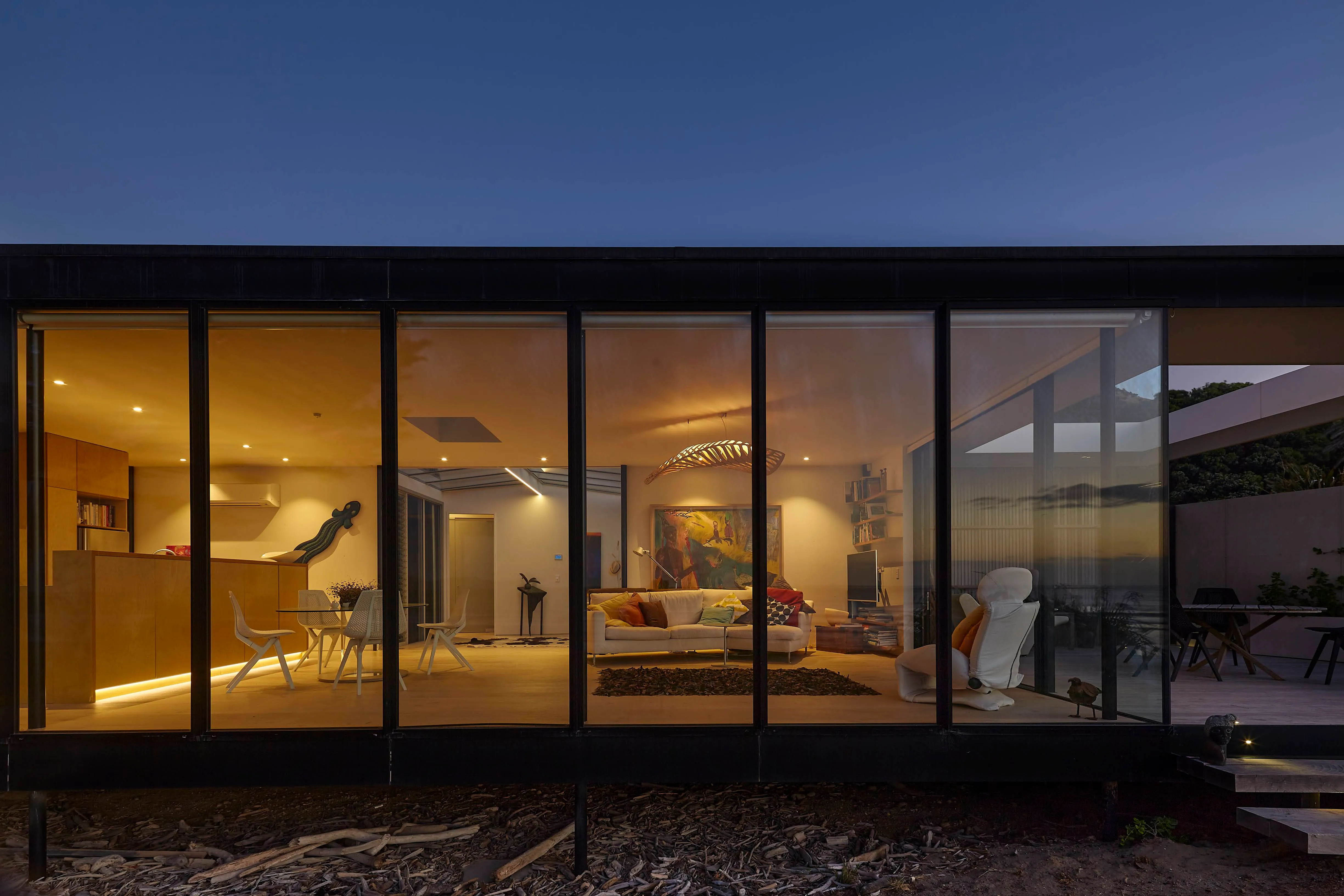 The completed beach house at dusk, its long glazed elevation lit warmly from within, the black-clad volume raised on slender posts above a bed of driftwood.