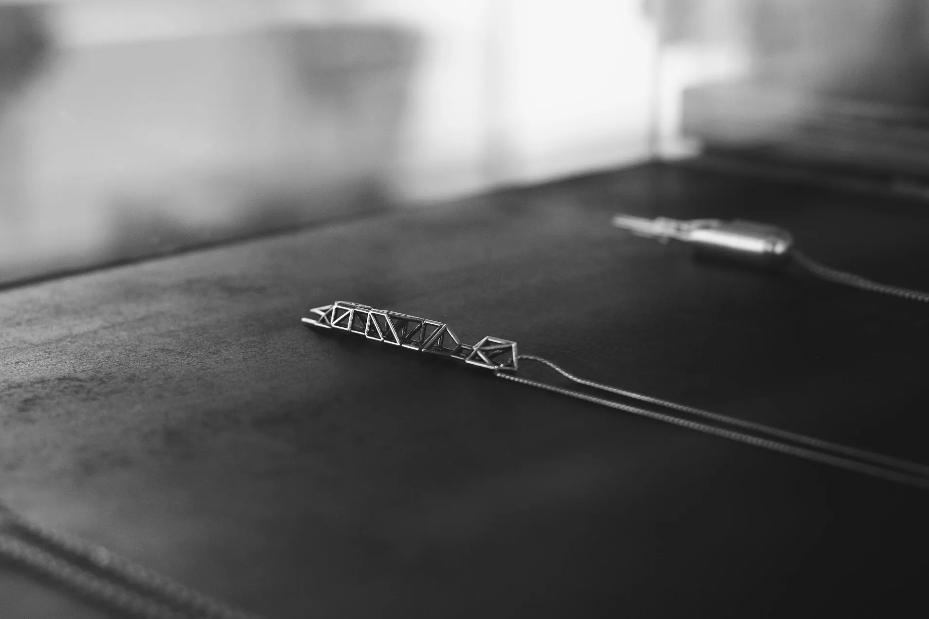 A slender silver truss pendant lying on a dark leather desk, its chain running out of frame toward a second pendant behind it.