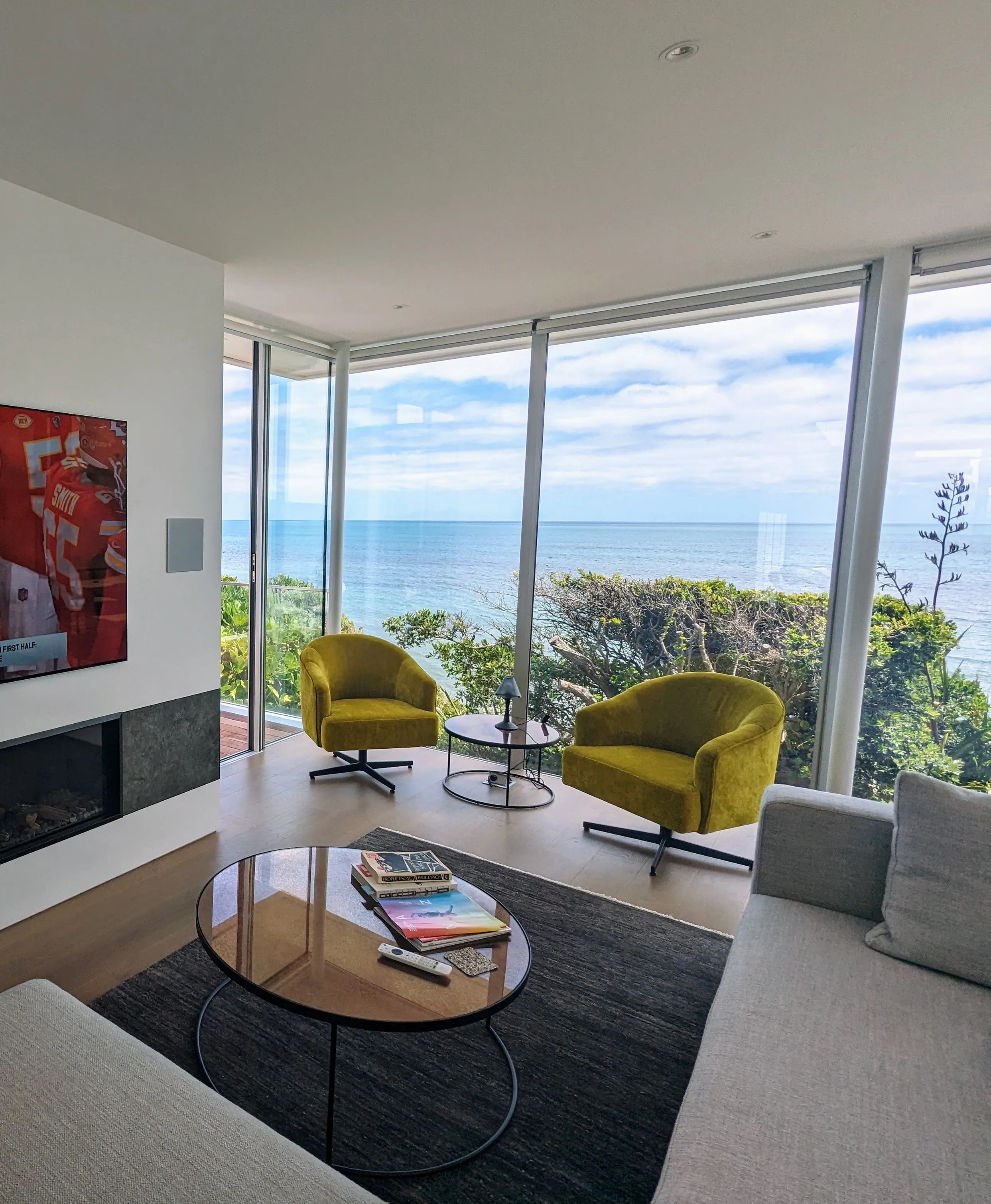 Completed living room looking out through a glazed corner to the sea, with two yellow armchairs at the window and a long linear fireplace set into stone on the left.