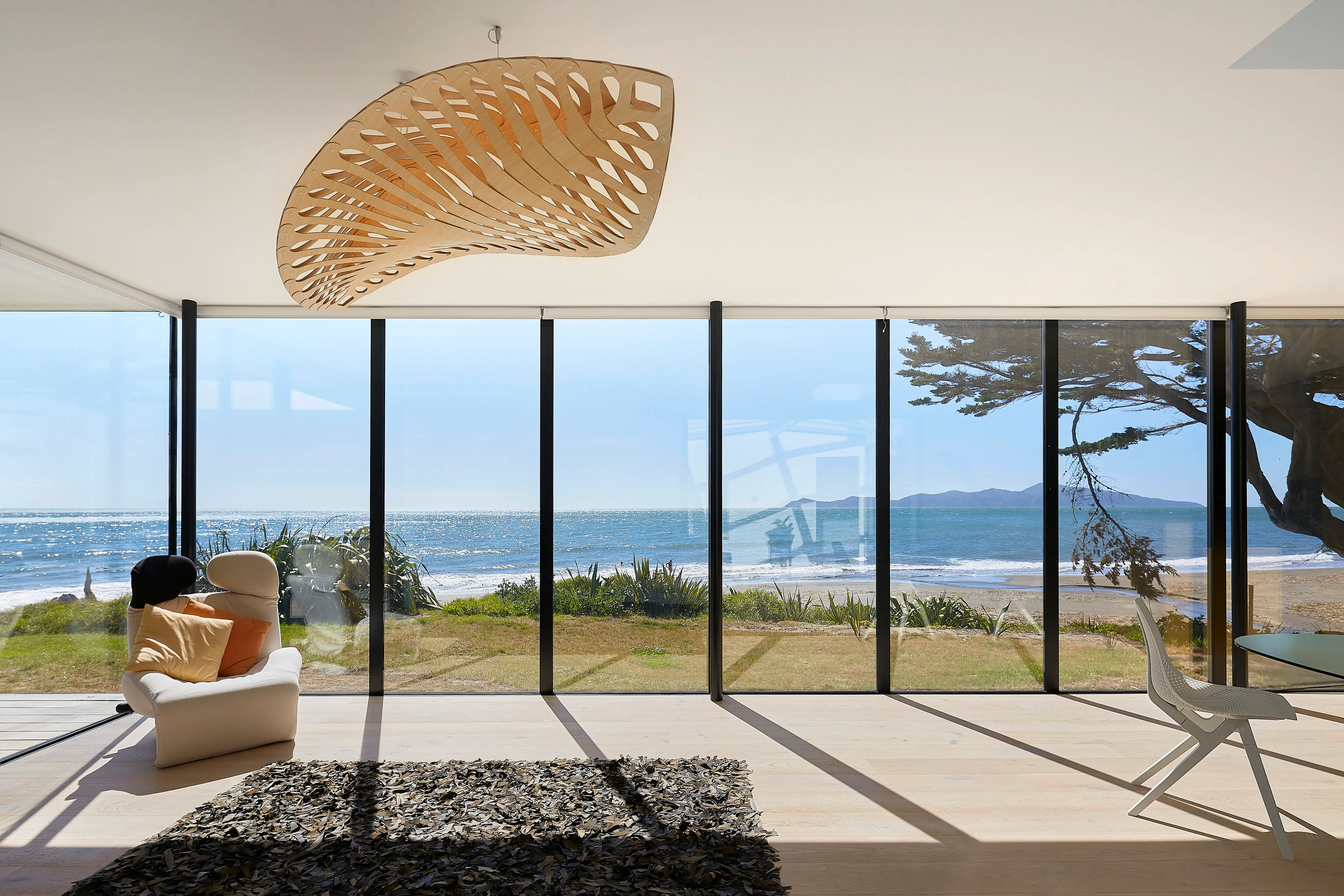 Living space behind full-height glazing looking out to the sea and a distant island, with a carved timber pendant light suspended from the ceiling.