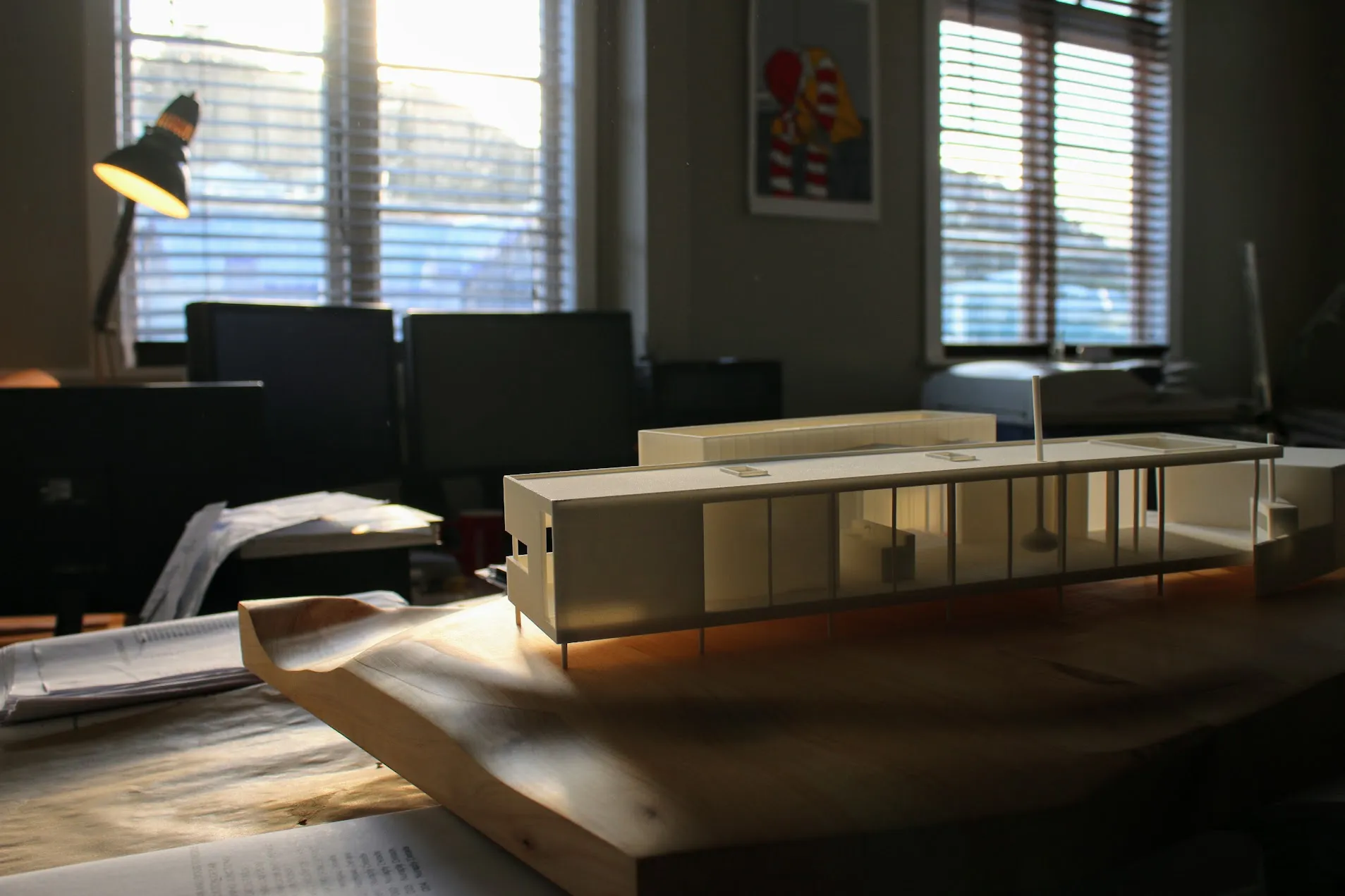 Large white 3D printed architectural model of the house raised on slender columns, photographed on a timber desk in an office with afternoon light behind it.