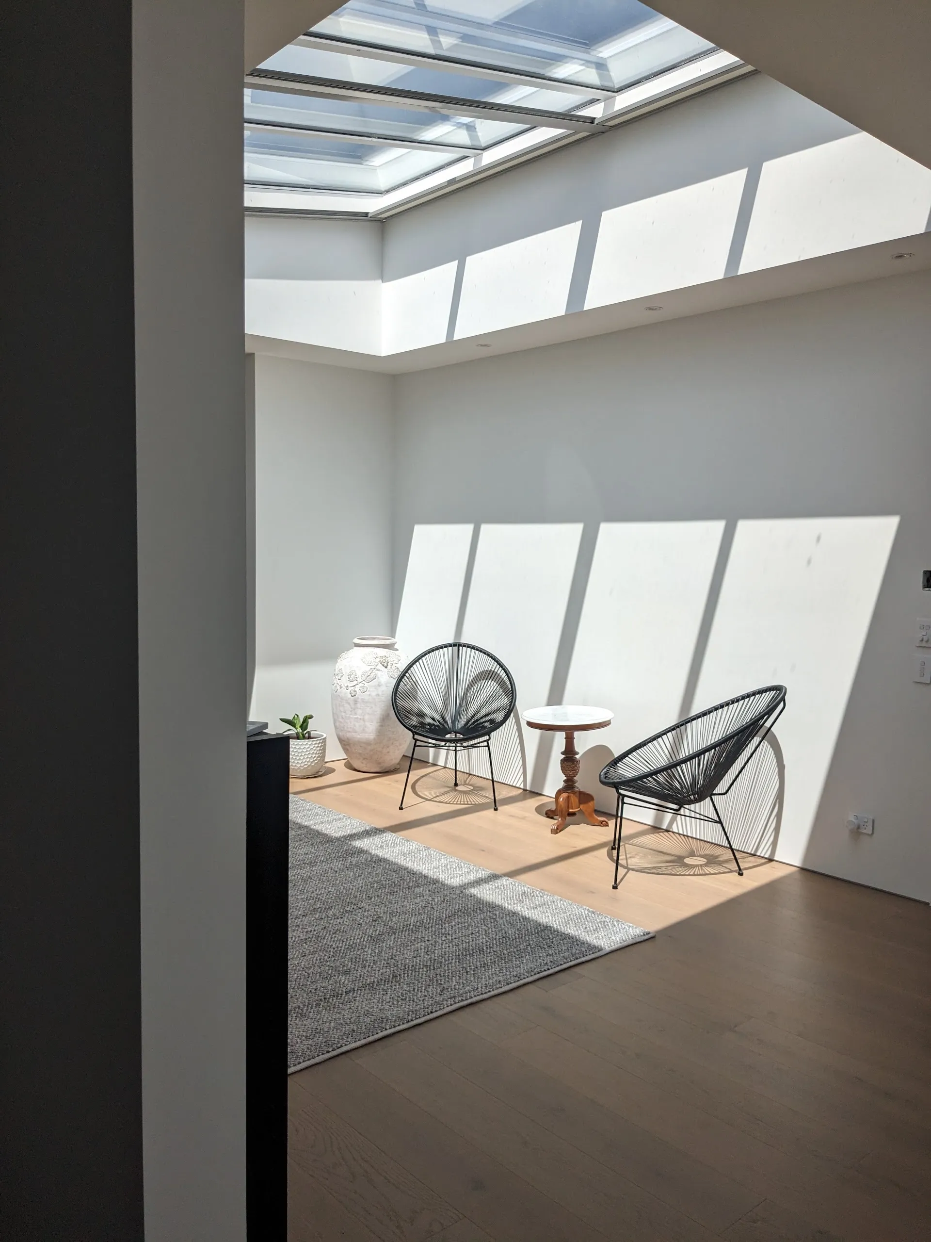 Completed interior seating area where a long roof lantern casts sharp rectangles of daylight down a white wall behind two black wire chairs.
